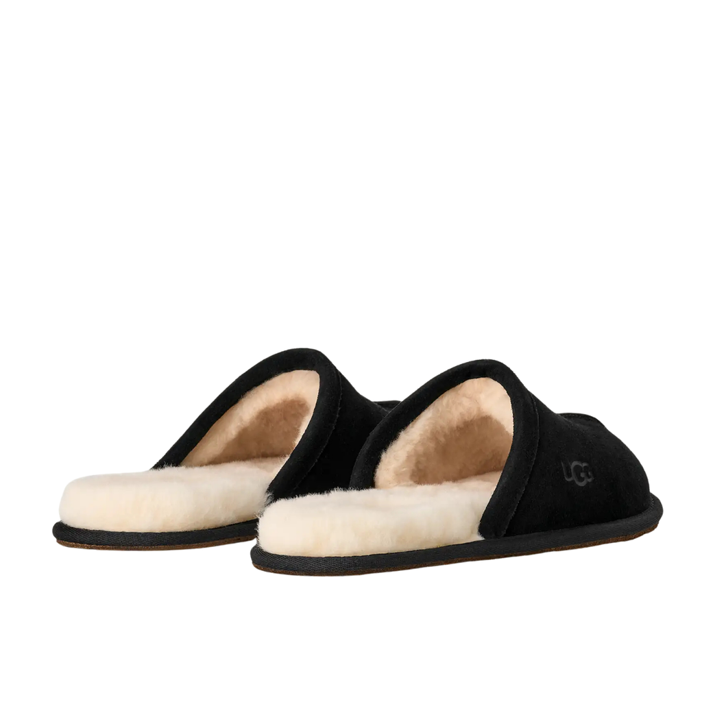 A pair of black slip-on slippers with a soft cream-colored shearling lining, shown from a side profile view on a plain white background. A small UGG logo is visible on the side of the right slipper.